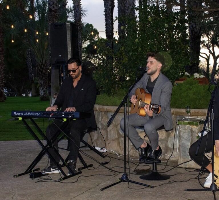 Dos músicos con guitarra española, micrófono y teclado en un jardín.